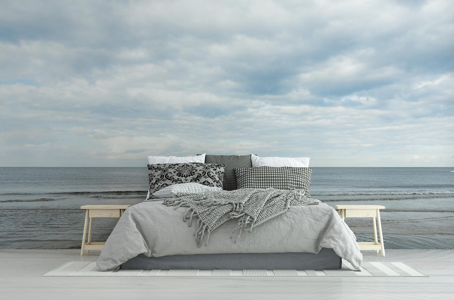 Bett mit grauer Decke, gemusterten Kissen und grauem Plaid seitlich zwei Holzbänke vor Meer und bewölktem Himmel Tapete von Superwalls
