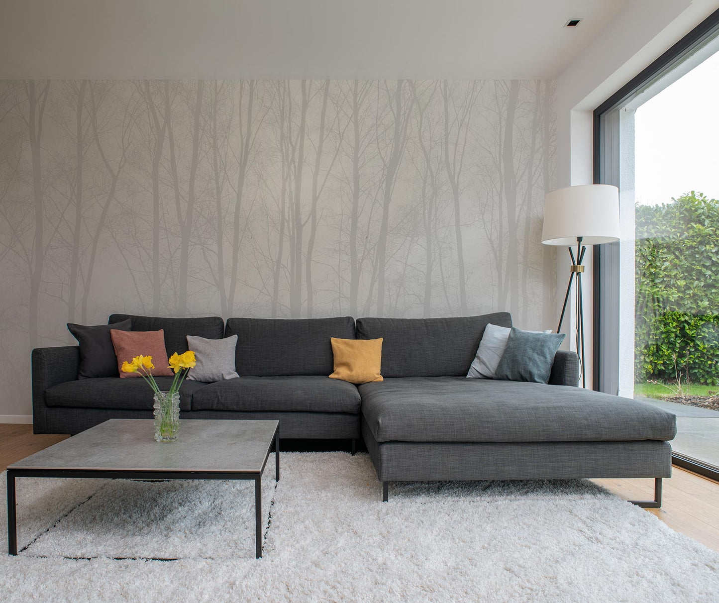 Dunkelgraues Ecksofa mit fünf bunten Kissen vor einer Wand mit Baumtapete von Superwalls, niedriger quadratischer Couchtisch mit Glasvase und gelben Blumen, großer Fensterblick auf Garten.