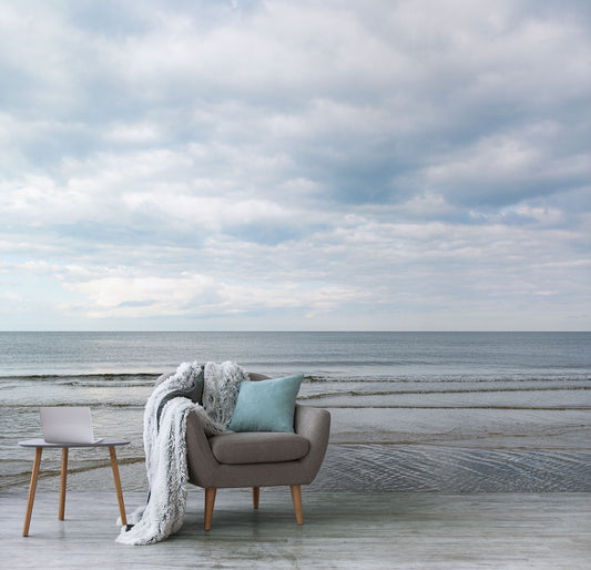 Beiger Sessel mit blauem Kissen und weißer Decke neben kleinem Holztisch mit Laptop vor einer Wand mit Meeresblick und bewölktem Himmel Tapete von Superwalls