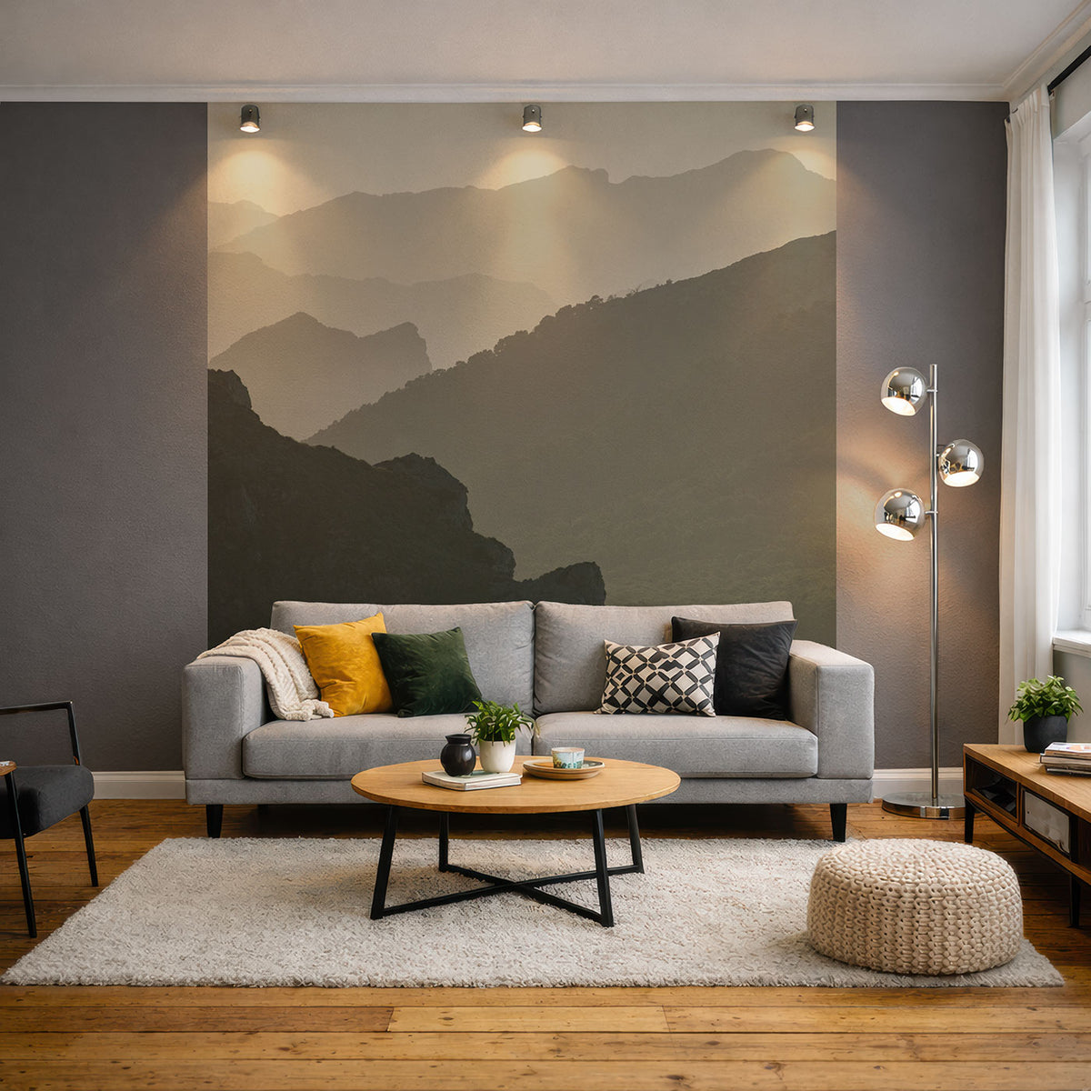 Wohnzimmer mit grauem Sofa, bunten Kissen, rundem Holztisch, Teppich, Pouf und Stehlampe vor Wand mit Berglandschaft-Tapete von Superwalls