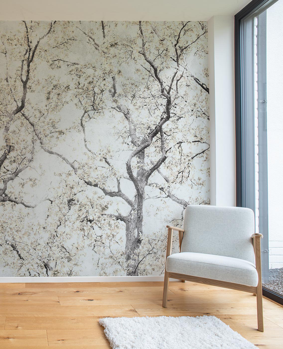 Hellgrauer Sessel mit Holzgestell vor einer Wand mit Baumzweig-Tapete von Superwalls neben bodentiefem Fenster und hellem Holzboden mit weißem Teppich