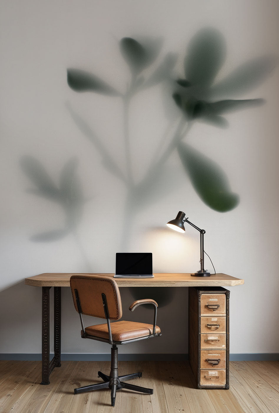 Schreibtisch mit Laptop, Schreibtischlampe und braunem Drehstuhl vor Fototapete mit großem unscharfem Pflanzenschatten