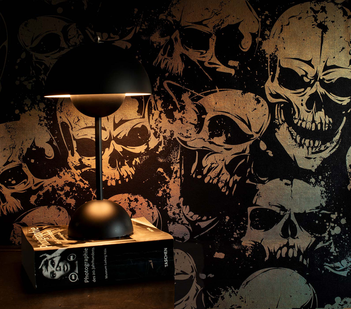 Schwarze Tischlampe mit halbkugelförmigem Schirm auf einem Buch vor einer Tapete mit großflächigem Totenkopf-Muster im metallic von Superwalls