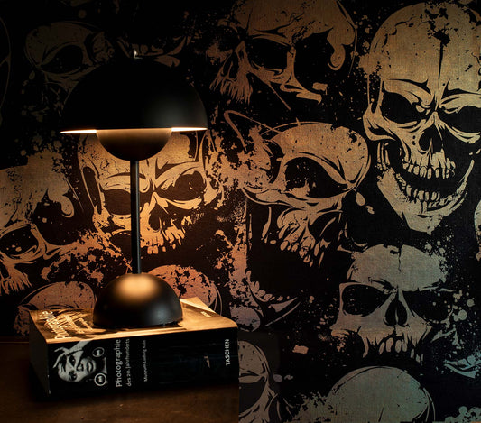 Schwarze Tischlampe mit halbkugelförmigem Schirm auf einem Buch vor einer Tapete mit großflächigem Totenkopf-Muster im metallic von Superwalls